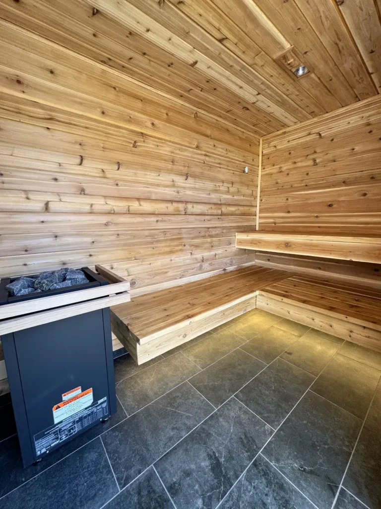 Western red cedar sauna interior with two-tier L-shaped benches, Harvia heater with stones, and slate tile floor