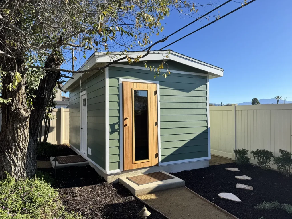 Custom backyard sauna with sage green Hardie lap siding, white trim and a cedar sauna door