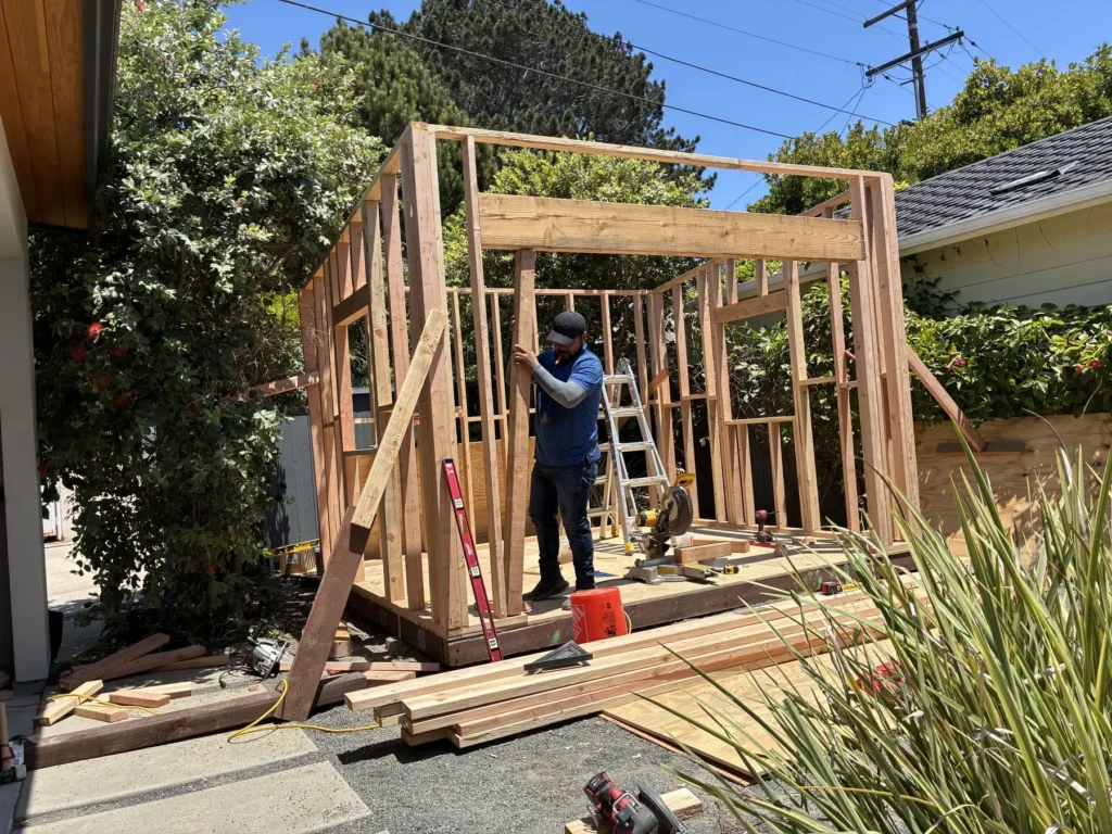 Builder framing the walls of a custom backyard building on site with lumber staged on the ground